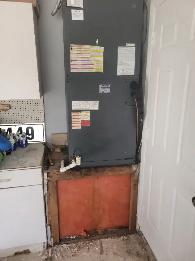 Existing air handler in a garage sitting on a water-damaged wooden platform with exposed, stained insulation and crumbling drywall at the base.