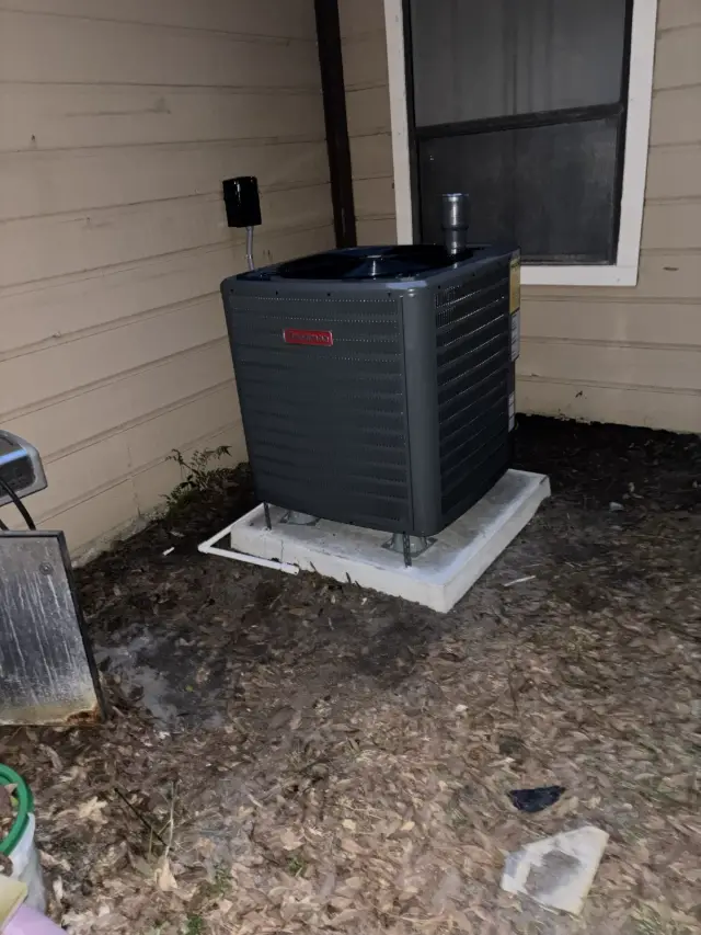 Goodman condenser installed on a new concrete pad against the side of a home, with an electrical disconnect above it and PVC condensate line routed along the ground.