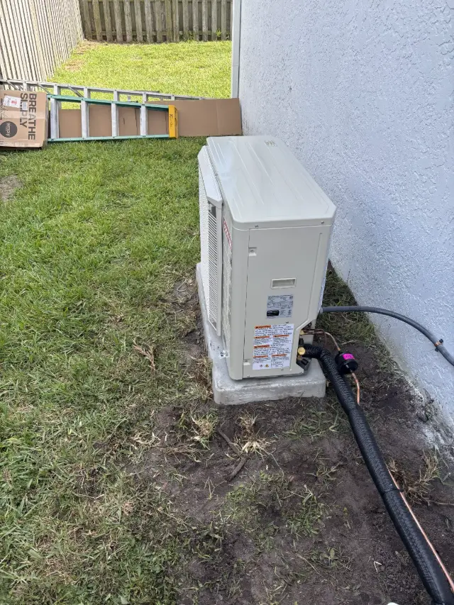 Rear view of a newly installed Goodman R32 condenser on a concrete pad in a grass yard, showing the insulated line set and service valves.