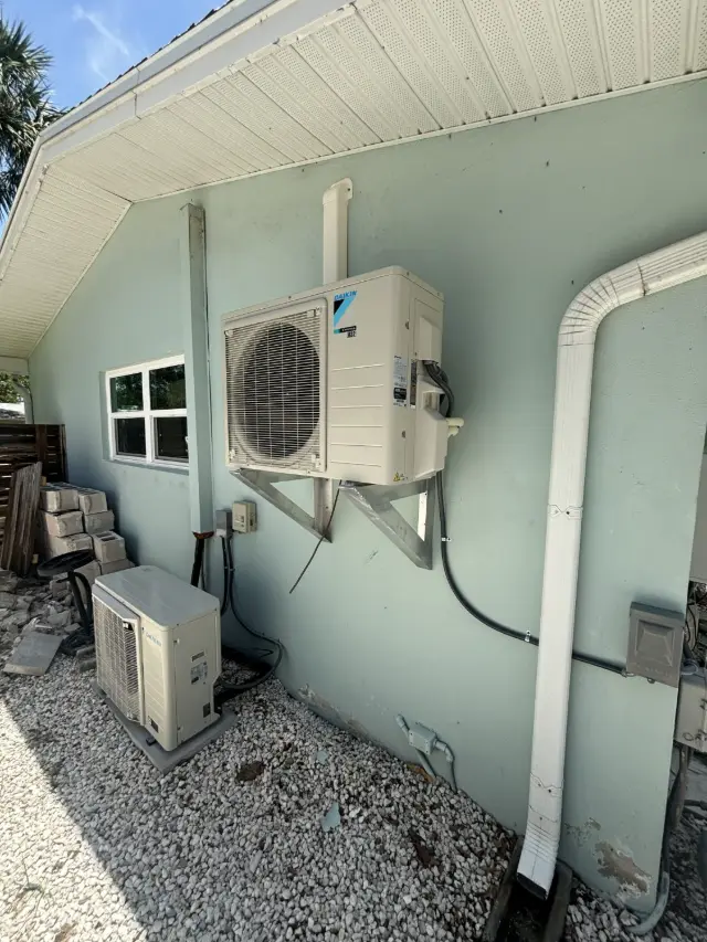 Two Daikin mini-split condensers outside a home, one mounted on a stainless wall bracket and one on a ground pad, with line sets neatly secured to the wall.