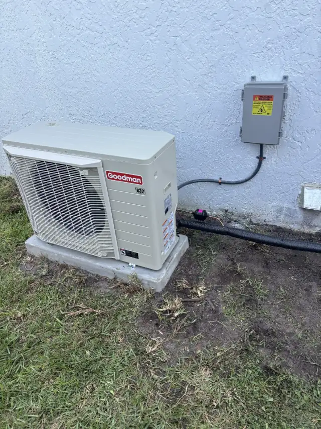 Newly installed Goodman R32 side-discharge condenser on a concrete pad beside a stucco home, with a weatherproof electrical disconnect mounted on the wall.