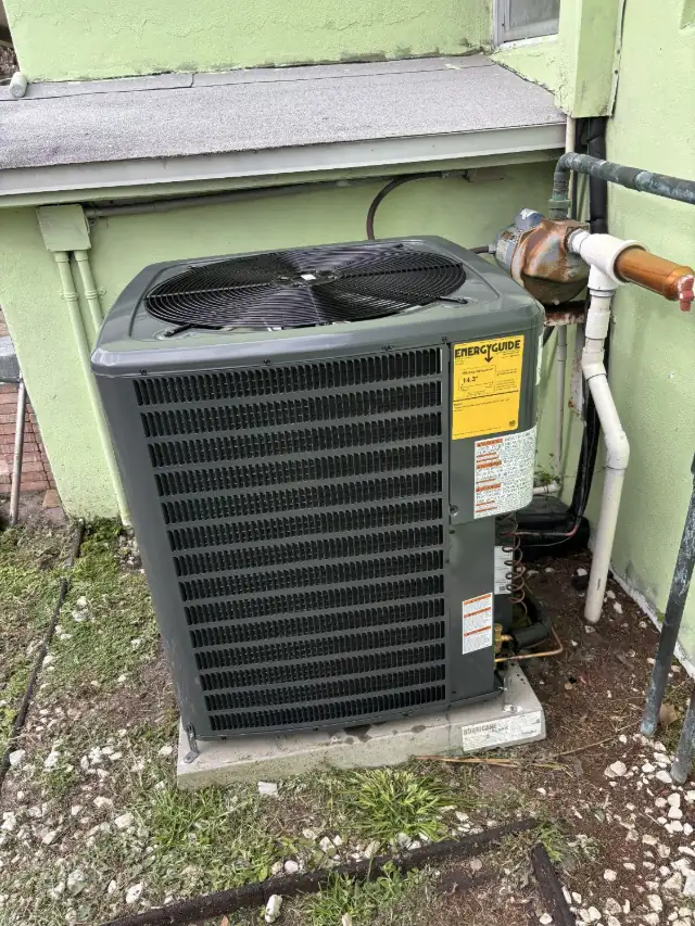 Goodman condenser on a hurricane-rated pad beside a green stucco home, with the copper line set and irrigation piping alongside.